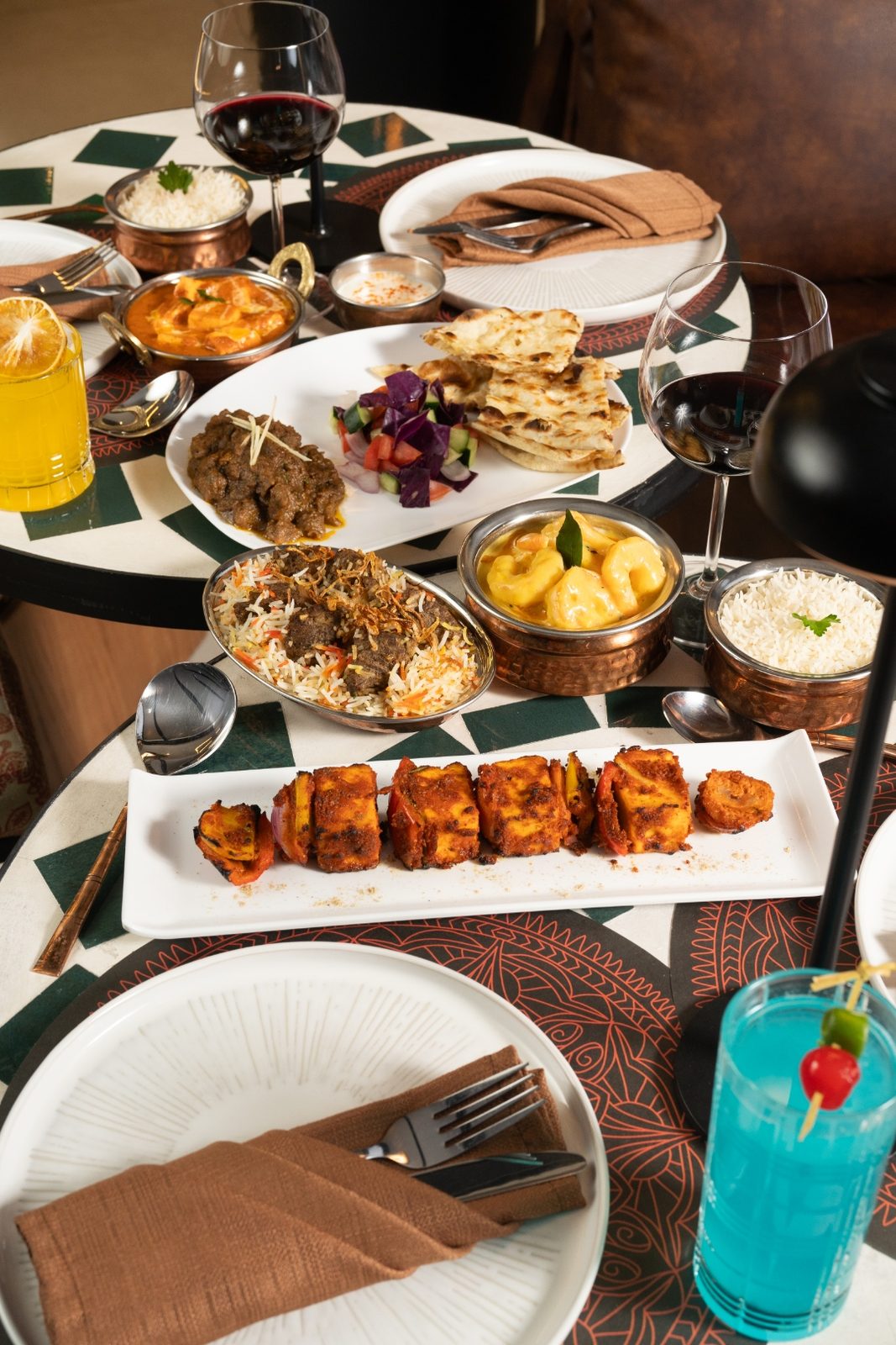 A rich spread of real Mantraa dishes on the table — lamb chops, biryani, paneer tikka, curries, naan, wine, and a cocktail