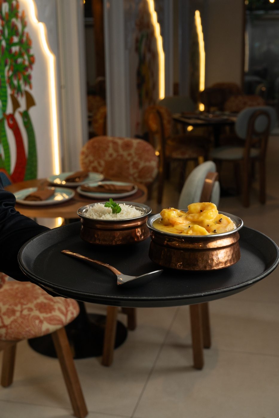 A server carrying a tray of prawn curry and rice through Mantraa's warm, moody dining room with a colorful mural
